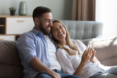 Güzel bir çift akıllı telefonlarıyla kanepede dinleniyor, internette eğlenceli videolar izlerken gülüyorlar. E-alışveriş, mobil uygulamaların mutlu müşterileri modern teknoloji, eğlence konsepti kullanarak kullanım, eğlence, konfor hayatı
