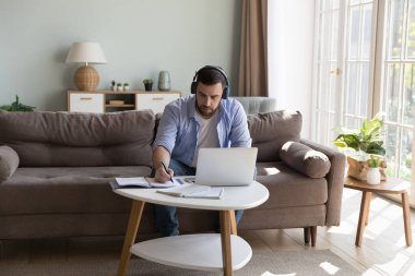 Kulaklıklı ciddi bir adam evden çalışır, işi çözer, video konferans uygulamasını laptopta kullanır, notlar alır, not tutar, internetteki öğretmenle dili geliştirir, eğitim, teknoloji konsepti