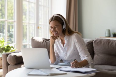 Kulaklıklı kadın video uygulaması kullanarak evden ders çalışıyor, dizüstü bilgisayarla masaya oturuyor, internetteki öğretmenle İngilizce bilgisini geliştiriyor. Uzak sınıf, yeni beceriler, sanal toplantı konsepti