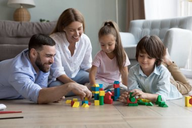Küçük ebeveynler ve küçük çocuklar çok renkli tahta tuğlalar oynuyorlar, oturma odasında sıcak bir zeminde oturuyorlar, haftasonu eğlencesini modern dairede geçiriyorlar, oyun ve iletişimin tadını çıkarıyorlar. Çocuk gelişimi, eğlence konsepti