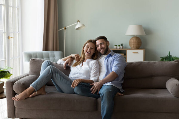 Beautiful millennial couple in love sit on sofa staring at camera. Concept of well-being family, homeowners spouses enjoy relaxation at new modern flat, independence, bank loan, romantic relationships