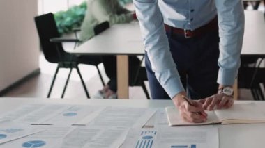 Ofiste çalışan bir işadamı, bilgi tablosuyla masaya yaslanmış, finansal verileri analiz ediyor, grafik ve diyagramlarla kağıtları gözden geçiriyor, notlar alıyor, strateji geliştiriyor, evrak işleri yapıyor, kırpılmış görüşü kapatıyor.