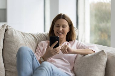 Modern akıllı telefonuyla rahat bir kanepeye uzanan, e-randevu hizmetlerini kullanan, arama yapan, sosyal medyadan keyif alan, boş zamanlarını evde yalnız geçiren güzel bekar bir kadın. Kablosuz teknoloji, uygulamalar.