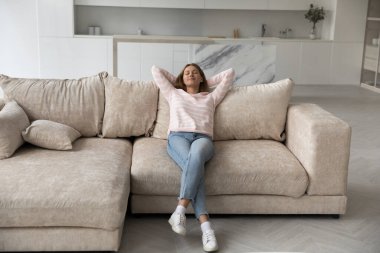 Uzun, huzurlu bir kadın, gündelik giysiler içinde kanepeye yaslanmış, evde tek başına dinleniyor, kadın moladan ve modern evde temiz havadan hoşlanıyor, stressiz bir hafta sonu geçiriyor, kendi evinde keyifli bir gün geçiriyor.