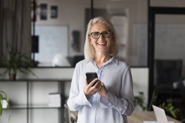 İş yerinde akıllı telefonuyla poz veren, gülümseyen, kameraya bakan, internetten iyi haberler okuyan, yeni mobil iş uygulamalarının keyfini çıkaran, yeni uygulamalar kullanan orta yaşlı bir iş kadını.