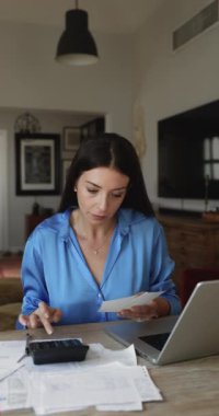 Oturma odasında oturan genç bir kadın ev faturalarını hesaplar, aile bütçesini yönetir, kişisel mali durumları analiz eder, harcamaları gözden geçirir, gelir ve sonuçları değerlendirir.