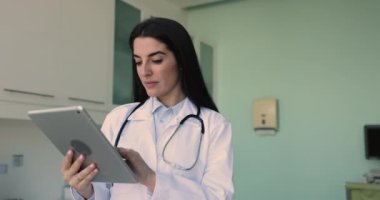 Ciddi genç İspanyol pratisyen kadın klinikteki dijital tablette interneti inceliyor, iş arkadaşı, hasta, online tıbbi tele-sağlık uygulaması kullanarak sohbet ediyor, mutlu oluyor.