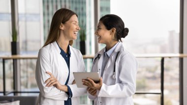 Young Indian and Caucasian women clinic staff members standing in office with tablet, laughing while lead informal conversation during break, share good news, having friendly relations at workplace clipart