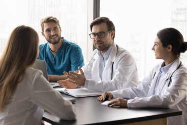 Sağlık uzmanlarından oluşan bir grup, tıbbi danışmanlık toplantısına katılmış, konferans masasına oturmuş, özel hasta vakalarını tartışıyor, genç takım liderinin konuşmasını dinliyor.