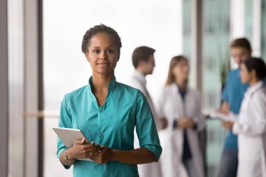 Modern klinikte poz veren turkuaz önlük giyen güzel Afrikalı kadın doktor portresi, kameraya bakıyor, dijital tablet tutuyor, arka planda bulanıklaşıyor. Görev, sağlık hizmetleri