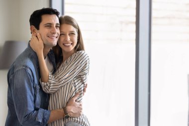 Mutlu, genç, evli bir çift, arka planda pencere ve gün ışığıyla kucaklaşıp fotokopi odasına bakıyor, gülümsüyor ve sıradan bir portre için poz veriyor.