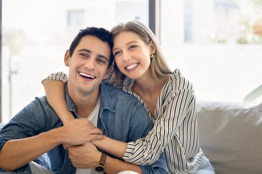 Neşeli genç İspanyol karı ve koca sevgiyle sarılıyorlar, kanepeye yakın oturuyorlar, başka tarafa bakıyorlar, gülümsüyorlar, gülüyorlar. Aşık mutlu çift romantik ilişkide mutluluğun tadını çıkarıyor.