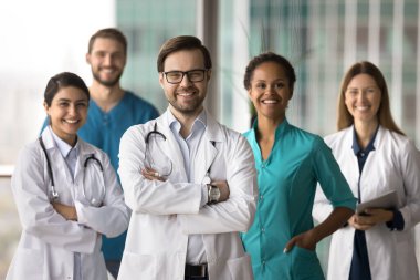 Erkek doktor kollarını kavuşturarak etnik çeşitliliğe sahip bir grubun önünde duruyor, gülümsüyor, birlikte kameraya bakıyor, modern klinik ofisinde poz veriyor. Hastane personeli portre, tıp