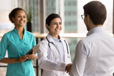 Mutlu Hintli kadın doktor beyaz önlüklü bir hasta ile Afrikalı bir hemşire karşılıyor, gülümsüyor, el sıkışıyor, klinikteki randevusuna başlıyor. Profesyonel sağlık hizmeti, tedavi, danışma