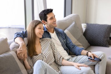 Evde birlikte romantik filmler izleyen, rahat bir kanepede dinlenen, sarılan, konuşan, gülen, uzaktan kumanda kullanan çekici genç bir çift.