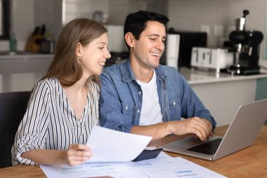 Finansal olarak başarılı genç çift kârı sayıyor, gelir analizlerini kontrol ediyor, dizüstü bilgisayardaki banka başvurusunu kullanıyor, faturaları kontrol ediyor, ödeme için kağıt faturalarını kontrol ediyor, gülümsüyor, gülüyor.