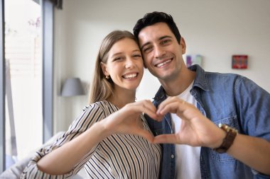 Mutlu genç çift el ele kalp hareketi yapıyor, sevgi, ilgi, şefkat, romantizm sembolü oluşturmak için parmaklarını birleştiriyor, kameraya bakıyor, portre için gülümsüyor.