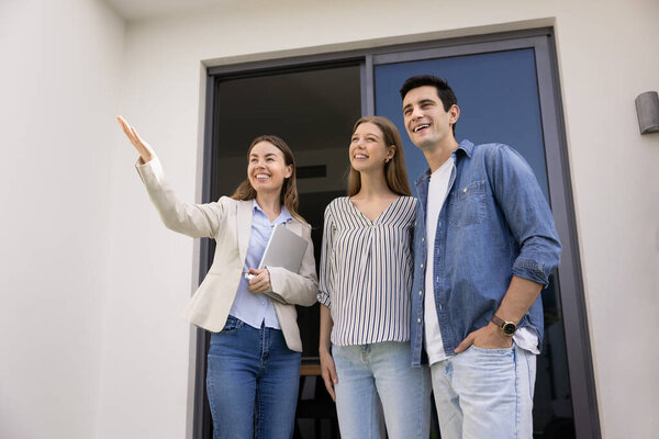 Positive realtor woman showing backyard, view from terrace to young couple of clients, renters, buyers, standing at doorstep at entrance to rental accommodation, pointing hand, looking away