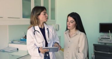 Genç İspanyol doktor kadın hastaya tablet ekranı gösteriyor, ultrason sonografi sonuçlarını açıklıyor, danışmanlık yapıyor, tedavi önerisinde bulunuyor.