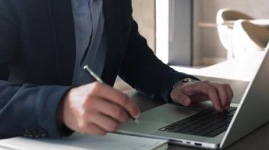 Takım elbiseli bir işadamı dizüstü bilgisayarında çalışıyor, elinde kalem tutuyor, not alıyor, kişisel organizatördeki önemli bilgileri not alıyor, planlama stratejisi, e-öğrenme, yeni bilgiler kazanıyor, görüşü kapatıyor.
