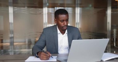 Ciddi Afrikalı iş adamı takım elbiseli, ofiste çalışıyor, bilgi araştırıyor, kağıda not alıyor ve dizüstü bilgisayar kullanıyor, kontrol raporları yazıyor, internette web sörfü bilgileri yazıyor, önemli verileri karalıyor.