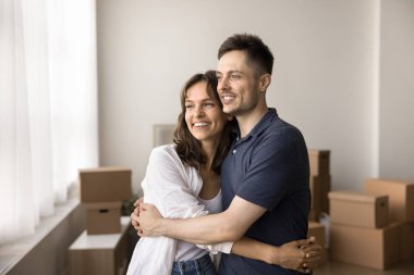 Mutlu ev sahipleri ailesi, kucaklaşan genç çift hoş düşünceler ve hayallerle dolu pencereden dışarı bakıyor, taşınma gününde dinleniyor, arka planda eşyalar olan karton kutular. Banka kredisi, ilk mülk