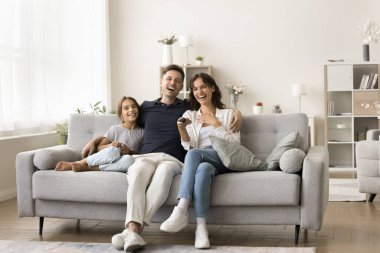 Aile üyeleri, genç ebeveynler ve tatlı küçük kız evde kanepede oturur, kanalı değiştirir, komedi filmi veya eğitim programı izler, paylaşılan aktivitelerin, birlikteliğin ve kaliteli zaman geçirmenin tadını çıkarırlar.