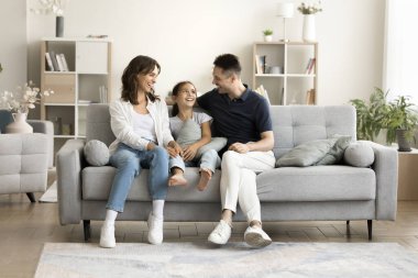 Küçük kız, sevgi dolu anne babasıyla kanepede oturur, gülümser, sohbet eder, hafta sonu tatili planlar, evdeki modern oturma odasında birlikte vakit geçirir. Aile bağları, güven, iletişim, anlayış