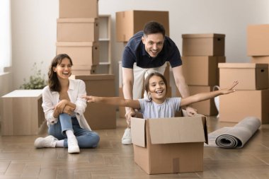 Heyecanlı aile taşınma gününde oyun oynuyor, baba küçük kızıyla birlikte karton kutuyu itiyor, mola veriyor, yeni eve taşınırken eğleniyor, mutluluk ve neşe saçıyor.