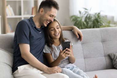 Küçük kız ve genç baba koltuğa oturup cep telefonu kullanıyor, komik videolar izliyor, video çağrı uygulaması ile ailesiyle uzaktan iletişim kurmanın keyfini çıkarıyorlar. Modern teknoloji, eğlence.