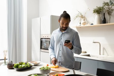 Ciddi genç siyah aşçı cep telefonundan yemek tarifi okuyor, danışmanlık yapıyor, web sitesi, organik gıda malzemelerinden yemek pişiriyor, salata için sebze kesiyor, online uygulama ile sipariş veriyor.