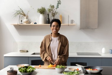 Mutlu Afrikalı şef kız ev mutfağında vejetaryen yemekleri pişiriyor, masada taze sebzeleri dilimliyor, organik doğal malzemeler hazırlıyor, yemek yapıyor, kameraya bakıyor, gülümsüyor.