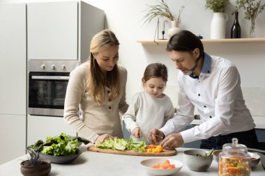 Odaklanmış küçük kız evlat anne ve babasına salata pişirmeleri için yardım ediyor, gemide taze sebzeler doğruyor. Ebeveynler ve tatlı kız mutfakta sağlıklı yemek için malzeme hazırlıyorlar. Aile sağlıklı yemek.
