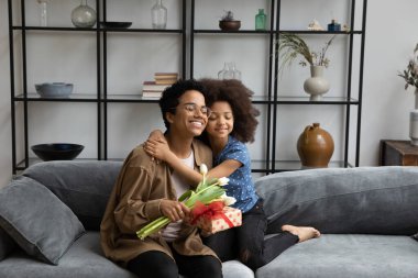 Genç Afrikalı mutlu bir çocuk. Minnettar bir anneyi kucaklıyor. Elinde hediye kutusu, lale çiçekleri, koltukta dinleniyor, gülümsüyor. Anne ve çocuk anneler gününü, doğum gününü, şenlik etkinliğini kutluyor.