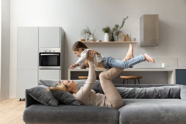 Mutlu aktif anne küçük kızını havaya kaldırıyor, rahat kanepeye uzanıyor, gülüyor, çocuklarla akroyoga yapıyor. Heyecanlı kız ve anne uçuyor, egzersiz oyunundan zevk alıyor.