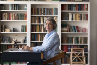 Ciddi bir son sınıf profesörü, kütüphanede dizüstü bilgisayarda çalışan olgun bir üniversite öğrencisi, arka planda kitaplıklar olan iş yerinden kameraya bakıyor. İçerideki portre