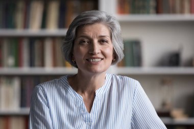 Olumlu, mutlu, yaşlı, iş öğretmeni kadın, vesikalık portresi. Dostça gülümseyen gri saçlı bayan kameraya bakıyor, arka planda kütüphane kitaplıklarıyla poz veriyor.