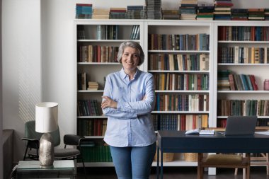 Kendinden emin yaşlı iş kadını, profesör, öğretmen portresi. Kütüphanede, ofis masasında kameraya gülümseyen, sıradan pozlar veren olgun bir kadın.