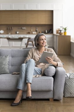 Yaşlılar ve modern teknoloji. Şık gülümseyen olgun kadın kendine güvenen dijital tablet kullanıcısı hafta sonu evdeki kanepede rahatla ecommerce web sitesinden ev yenileme için çevrimiçi alışverişler yap