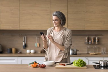 Akıllıca yemek. Kendine güvenen orta yaşlı kadın ev mutfağında doğal sebze yemeği pişirirken internette sağlıklı salata tarifi yemek fikrini bulmak için cep telefonu uygulaması kullanıyor. Boşluğu kopyala