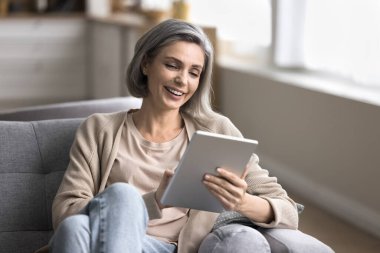 Sosyalleşme ve iletişim. Portre gülümseyen orta yaşlı kadın oturma odasındaki kanepede dinleniyor online öğrenim etkinliği uygulamalarına bağlı video görüşmesi ile dijital ped sohbeti yapıyor internet ödemeleri yapıyor