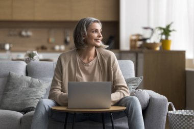 İş verimliliği ve ev içi rahatlığı. Mutlu olgun kadın serbest çalışan, rahat ev ofisinde dizüstü bilgisayarı kullan proje planı metni hazırla rahat koltukta otur konsept fikirden esinlenerek online bulundu