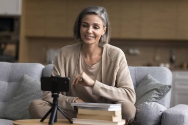Kitap incelemesi vlog. Kendine güvenen olgun kadın blogcu etkileyicisi video içeriğini ev ofis stüdyosundaki edebiyat bloguna üç ayaklı telefon kullanarak kaydeder kitap eleştirisi yapar yazarları seyirciye tavsiye eder
