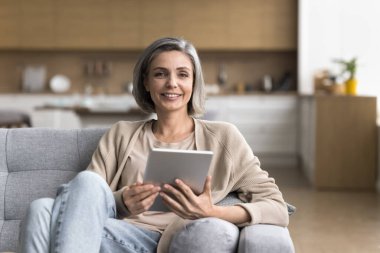 Dijital iletişim. Kendine güvenen, orta yaşlı, zarif bir kadının portresi rahat bir koltukta oturmuş elinde bilgisayarla kameraya bakıyor evden internetten video kursuyla öğreniyor.