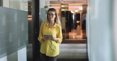 Kadın ofis koridorunda dijital tabletle dolaşıyor, çevrimiçi sohbeti yönetiyor, yazışıyor, e-posta yolluyor, veri analizi yapıyor, sunuma hazırlanıyor, güncellemeleri kontrol ediyor, çevrimiçi çalışıyor. Modern teknoloji kullanılarak çalışma akışı