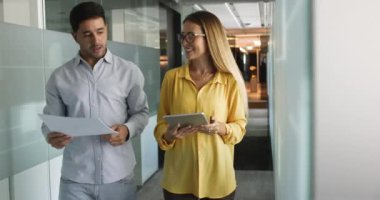 Ofiste koridorda yürürken konuşan iki iş arkadaşı, ellerinde kağıtlar ve dijital tablet, ortak proje üzerinde çalışıyorlar, güncellemeleri paylaşıyorlar, yaratıcı fikirler ve çözümler üretiyorlar.