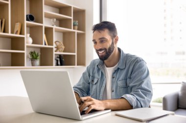 Genç gülümseyen İspanyol işadamı, öğrenci ya da ofis çalışanı öğrenim görüyor, e-öğreniyor, bilgi arıyor, yeni bir platform öğreniyor, elinde kalem tutuyor, modern bilgisayarla masaya oturmuş yararlı bilgiler yazıyor.