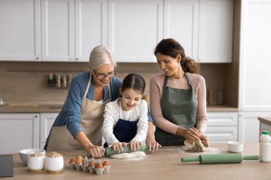 Family recipe. Intergenerational family older grandma young mum little child school age daughter grandkid cook at modern home kitchen together adult females teach kid knead roll dough for homemade pie clipart