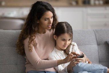 Ebeveyn kontrolü. Genç bekar anne, reşit olmayan kızıyla kanepeye oturup akıllı telefondaki eğitim uygulamasının kullanımına yardım ediyor. Elektronik sınıf kaydını indirerek notları tartışmayı öğrenme platformundaki görevi açıklıyor.