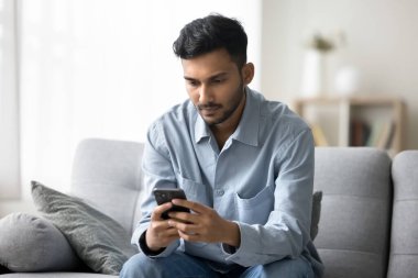 Konsantre genç adam serbest çalışıyor. Kanepede oturuyor. Cep telefonu kullanıyor. Sanal borsada döviz ticareti yapıyor.